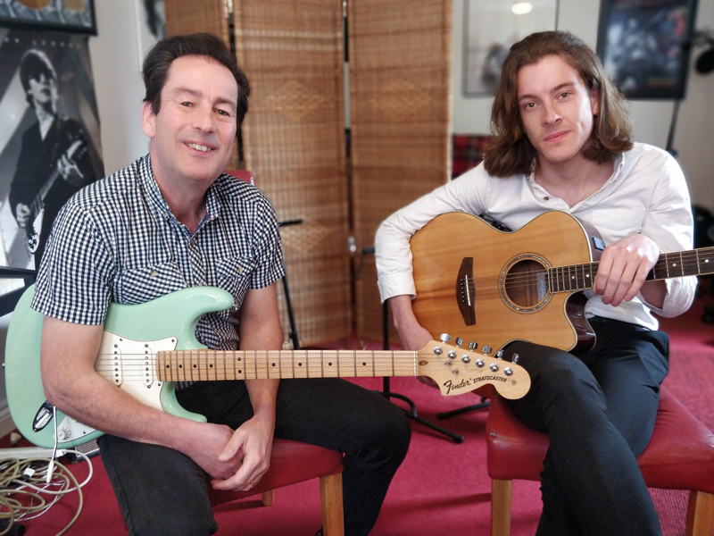 Paul and Mitch in their music studio holding an electric guitar and acoustic guitar.